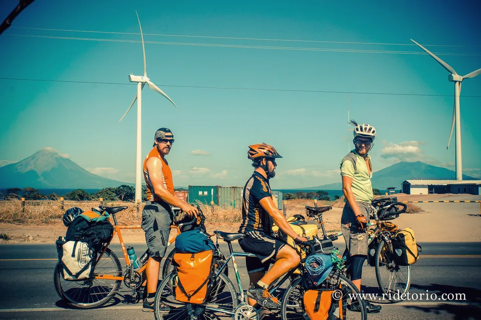 Three riders under a line of wind turbines, with a volcano across Lake Nicaragua.
