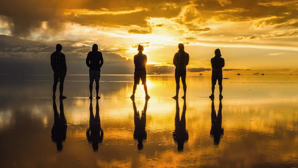 Five silhouettes stand on the mirror-flat salt at sunset, reflected perfectly beneath them.