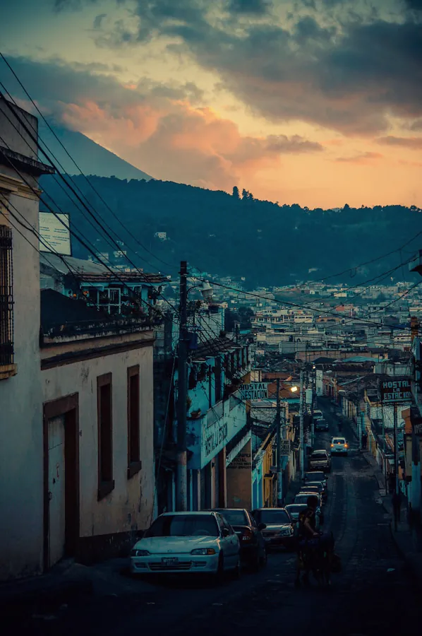 A colonial street at dusk in Xela with hostel signs and a volcano under orange clouds.